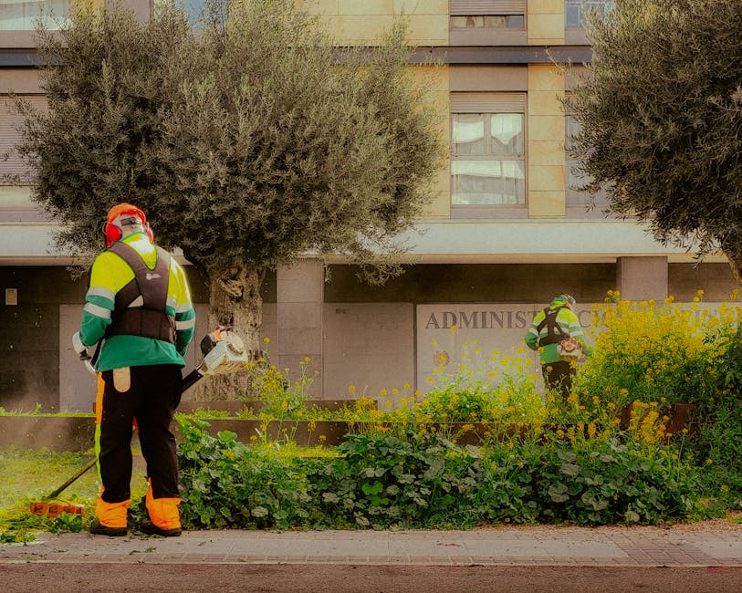 Two professional gardeners from gardenerkensington.co.uk are engaged in garden maintenance outside a modern building. One gardener stands in the foreground, holding a leaf blower and wearing high-visi